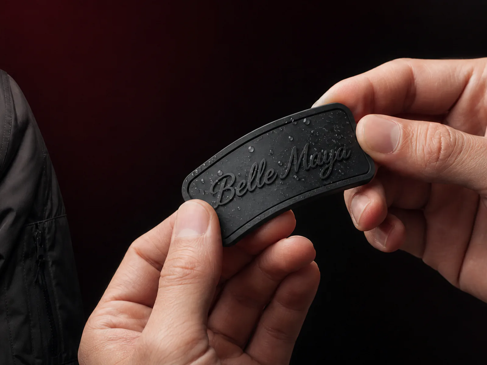 A maker flexing a wet black silicone patch with a raised Belle Maya wordmark beside a matching jacket patch